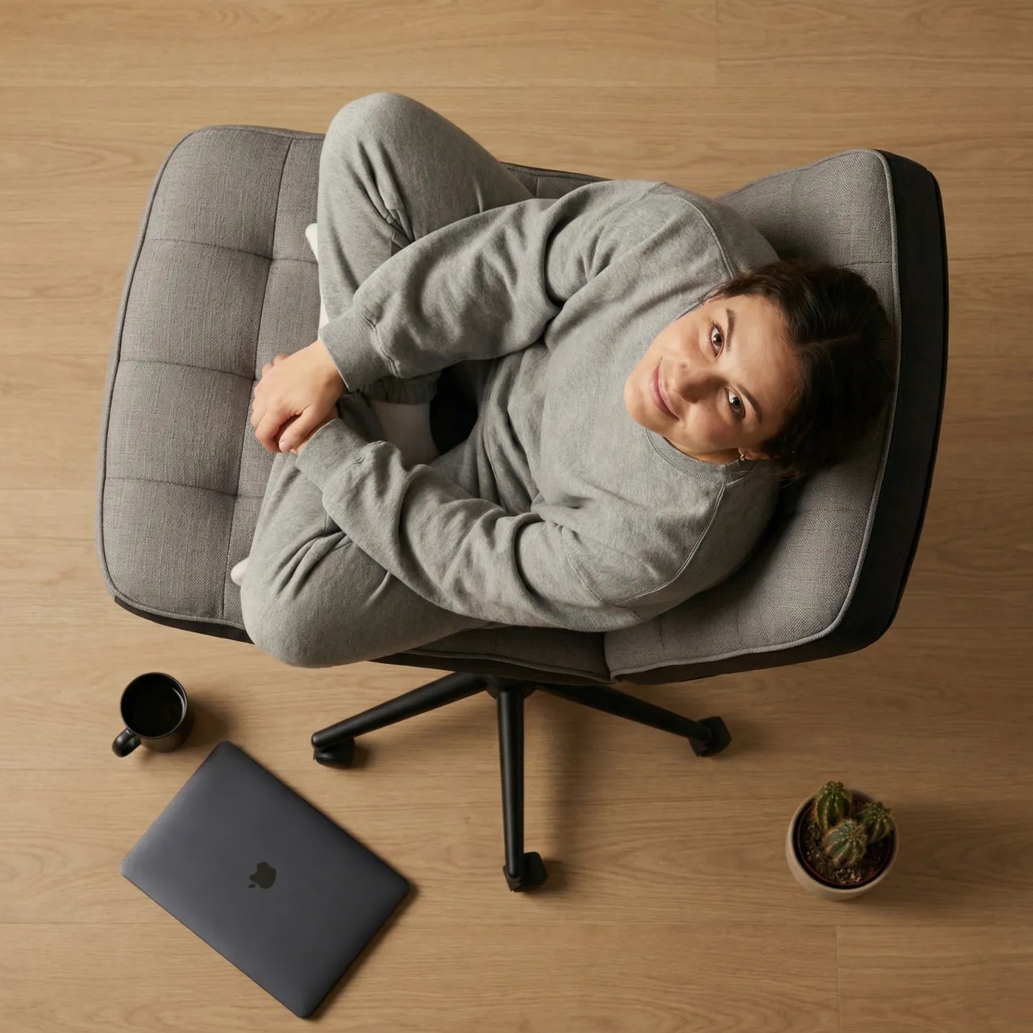 Vue aérienne femme détendue assise en tailleur sur la District 18 chaise bureau tissu lin gris capitonnée sans accoudoirs, parquet chêne laptop et cactus au sol
