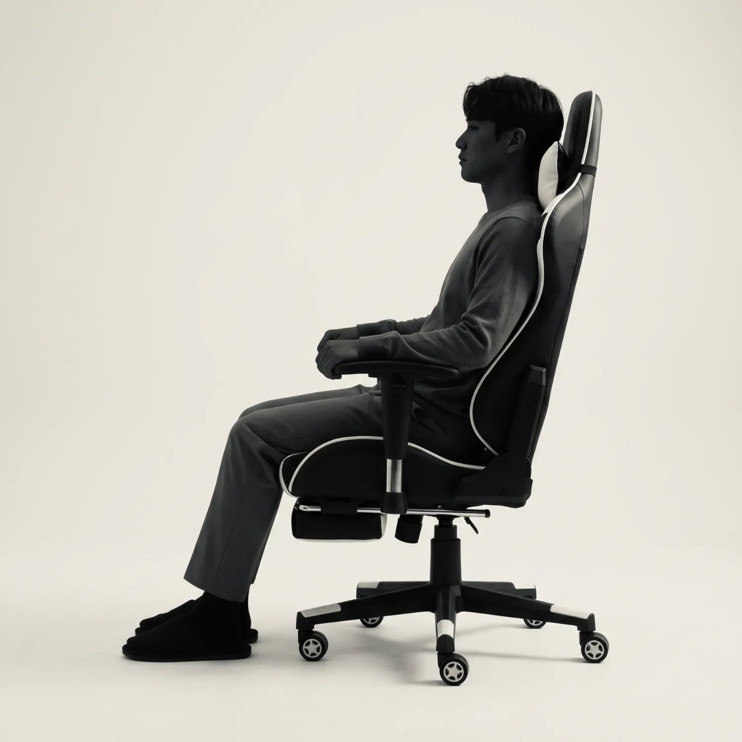 Homme assis de profil avec repose-pieds déployé sur la District 21 chaise gaming massage lombaire noir et blanc, posture gaming ergonomique fond studio neutre