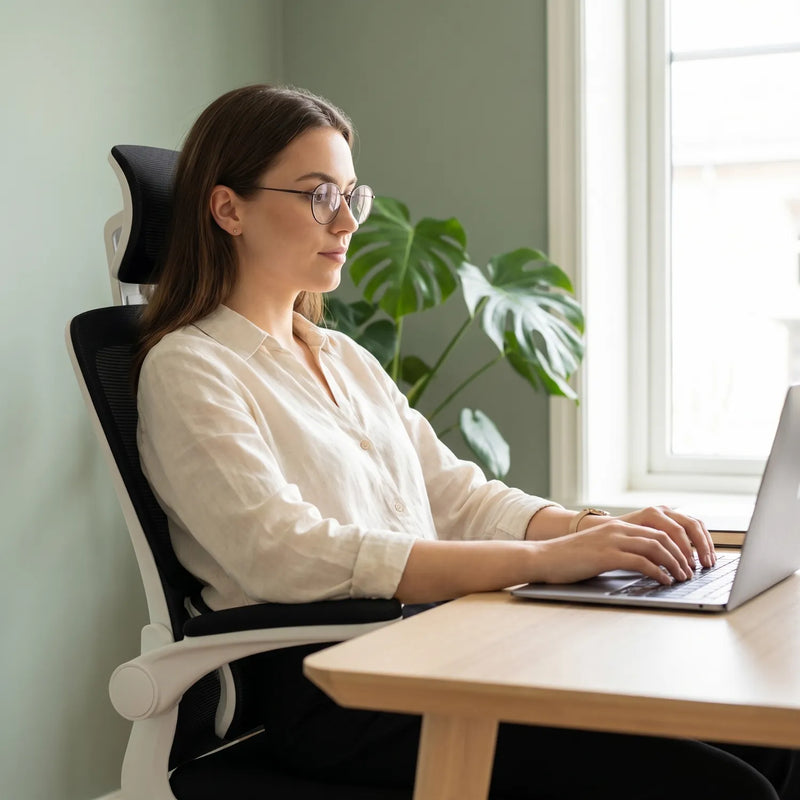 Femme avec lunettes en posture ergonomique sur la District 22 chaise bureau maille 3 segments noir et blanc, bureau bois clair lumière naturelle mur vert sauge monstera